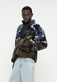 Geacă din fleece cu model camuflaj albastru, verde și maro, prevăzută cu guler înalt, închidere cu fermoar și două buzunare frontale. Textură moale.