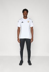 Fehér sportpóló fekete Adidas csíkokkal, klublogóval. Fekete nadrággal párosítva, amelyek fehér oldalsávokkal rendelkeznek, és fekete sportcipővel.