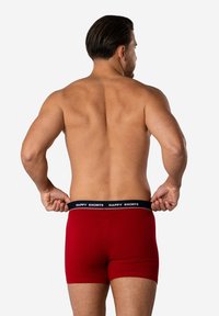 Červené boxerky s hladkou texturou a kontrastním černým pasem, na kterém je výrazně vytištěn text "ŠŤASTNÉ KRÁTKÉ".