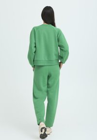 Zelená, nadrozmerná mikina a zodpovedajúce joggingové nohavice z jemnej látky, s ribovanými manžetami a pásom, zobrazené zozadu. Viditeľné tenisky.