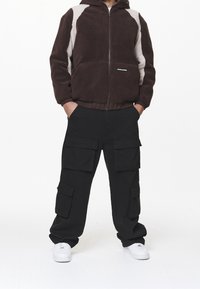 Felpa con zip marrone in pile con accenti grigio chiaro, abbinata a pantaloni cargo neri con grandi tasche e sneakers bianche.