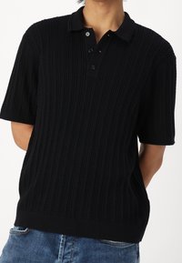 Polo negro de manga corta con un diseño texturizado acanalado, que presenta un collar clásico y una tapeta de tres botones. La tela parece suave.