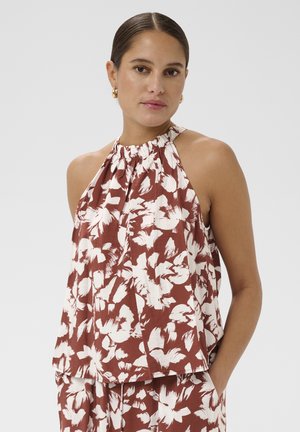 Femme portant un haut marron sans manches à col halter et un pantalon assorti avec de grandes impressions florales blanches, debout avec une main dans la poche.