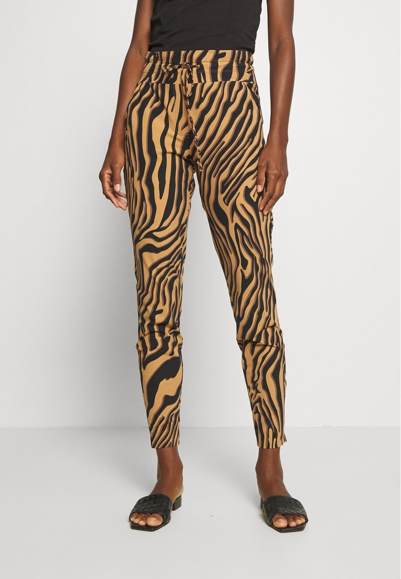 Pantalons rayés de zèbre marron et noir avec une coupe ajustée, une taille élastique et des poches latérales ; portés avec des sandales à glissière noires tissées.