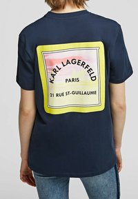 Tumma sininen t-paita, jossa on suuri keltainen graafinen kuva takana ja teksti "KARL LAGERFELD PARIS", sekä pehmeä ja sileä tekstuuri.