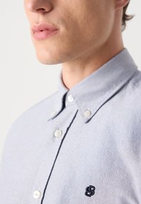 Lichtblauwe overhemddesign met een klassieke kraag, witte knopen en een klein geborduurd logo in marineblauw op de borst.