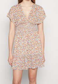 Robe fleurie avec un décolleté en V profond, des manches courtes évasées et une taille smockée. Comprend une jupe à volants dans des tons pastel avec des fleurs multicolores.