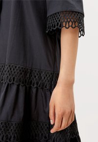 Černé šaty s vrstvovým designem a složitými krajkovými detaily. Krajka má geometrický vzor na rukávech a lemu.