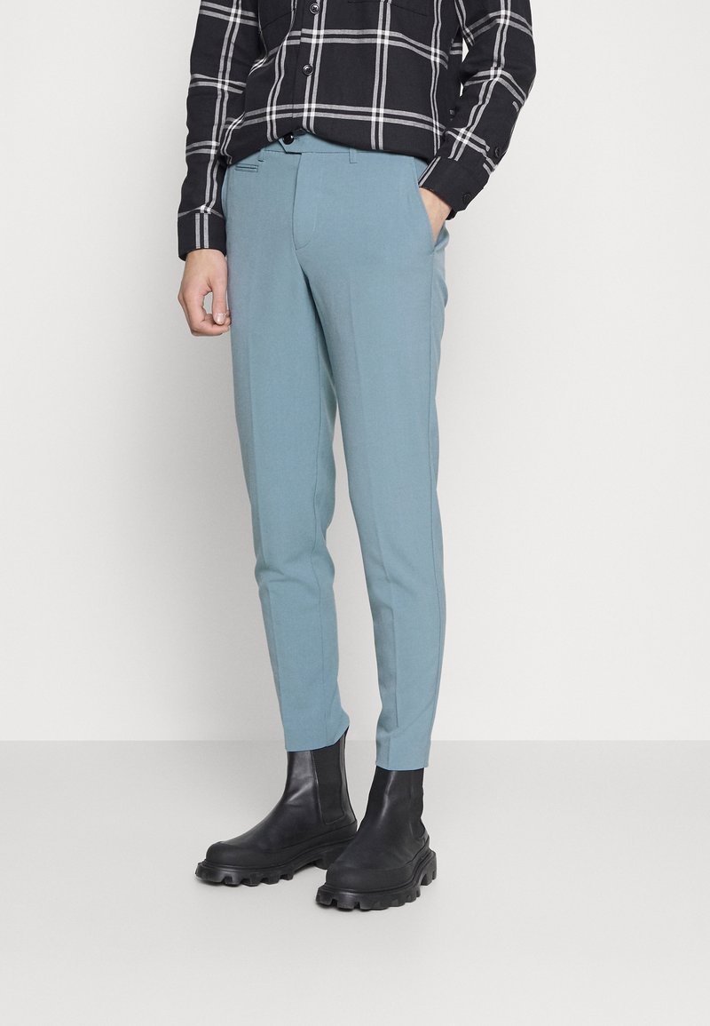 Světle modré slim-fit kalhoty z hladkého materiálu, s předními kapsami a záhybem, spárované s černými kotníkovými botami.