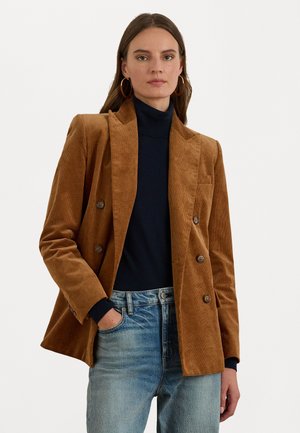 Blazer doppiopetto in velluto a corda marrone con revers a scollatura, tasche frontali e dettagli con bottoni. Indossato sopra un dolcevita nero e jeans blu.
