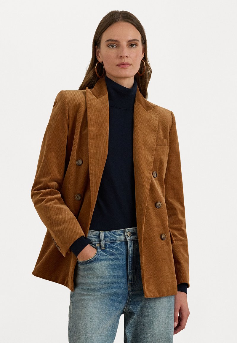 Bruine corduroy dubbelzijdige blazer met ingesneden revers, voorzakken en knoopaccenten. Draagt over een zwarte turtleneck en blauwe spijkerbroek.