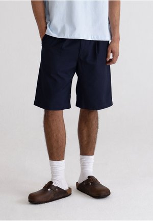 Mies pukeutunut laivastonsinisiin polven alle ulottuviin shortseihin, valkoisiin sukkiin, ruskeisiin liukukenkiin ja vaaleansiniseen paitaan, kädet taskuissa.