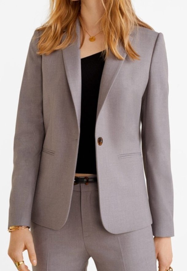 Femme portant un costume gris clair avec un blazer à un bouton sur un top noir, accessoirisée avec des bijoux dorés et une ceinture noire.