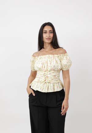 Femme aux longs cheveux foncés portant un haut style corset floral décolleté et un pantalon noir, debout avec une main dans la poche sur fond blanc.