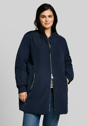 Frau mit dunklen Haaren, die eine lange marineblaue Reißverschlussjacke über blauen Jeans und einem gestreiften Oberteil trägt, steht vor einem schlichten grauen Hintergrund.