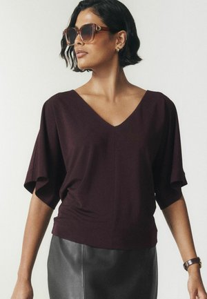 Femme aux cheveux courts et foncés portant de grandes lunettes de soleil marron, un haut violet foncé à col en V avec de larges manches, une jupe en cuir noire, des boucles d'oreilles dorées et une montre.