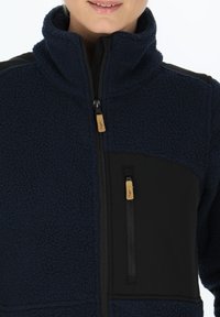Navy fleece jacket with a high collar, black panel accents, two zippered pockets, and a soft, textured fabric. Zipper pulls have wooden accents.