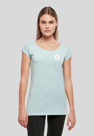 Jeune femme aux cheveux lisses porte un t-shirt bleu clair à manches courtes avec un petit motif de marguerite blanche sur la poitrine gauche.