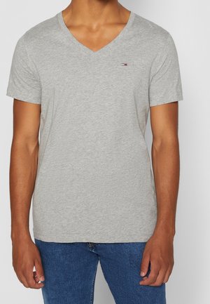 Mand iført en lys grå V-hals kortærmet T-shirt med et lille logo på venstre bryst og blå jeans, stående mod en ensfarvet baggrund.