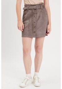 Mini-jupe en daim marron avec des boutons à l'avant, une ceinture avec un anneau en métal et une coupe droite. Associée à des baskets blanches et des chaussettes de cheville.