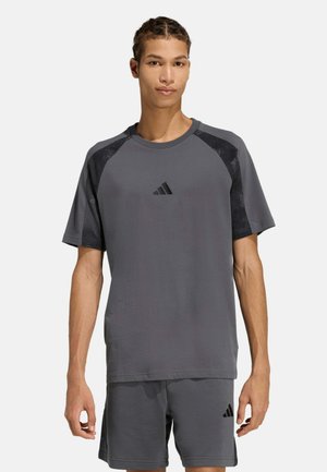 Joven con el pelo trenzado, vistiendo una camiseta deportiva gris de Adidas y pantalones cortos a juego, de pie frente a un fondo liso.