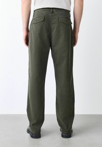 Man wearing olive green straight-leg pants with buttoned back pockets, black shoes, standing on gray floor against white wall.