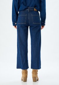 Jeans de pierna ancha en denim azul oscuro, con cintura alta, dos bolsillos traseros y dobladillos deshilachados. Combinados con botas de tobillo en color beige.