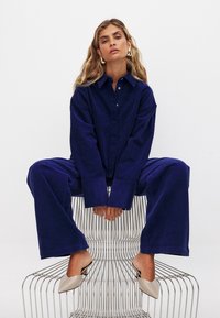 Tamsiai mėlynas kordūro dbutton-up marškinis derinamas su plačiomis kelnėmis, turinčiomis šonines kišenes ir tekstūrinį pavir�šių. Nuogas spalvos atviru kulnu bateliai užbaigia įvaizdį.