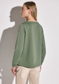 Zelená mikina s kulatým výstřihem a dlouhými rukávy, měkká textura, uvolněný střih a žebrované manžety, spárovaná s béžovými kalhotami. Stín vržený na zeď.