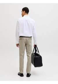 Άνδρας με λευκό πουκάμισο και μπεζ παντελόνι που κρατά μια μαύρη τσάντα duffel, στέκεται σε απλό φωτεινό φόντο, με την πλάτη γυρισμένη.