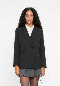 Kvinne med langt krøllete hår iført en mørkegrå blazer, hvit skjorte, grå plisserte skjørt og gjennomsiktige svarte strømpebukser, stående mot hvit bakgrunn.