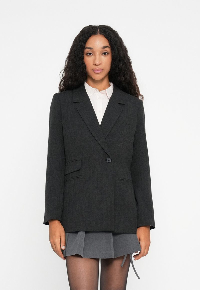 Kvinne med langt krøllete hår iført en mørkegrå blazer, hvit skjorte, grå plisserte skjørt og gjennomsiktige svarte strømpebukser, stående mot hvit bakgrunn.