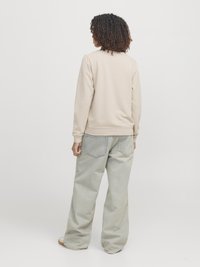 Kihara inimene, kes kannab beeži sweatshirt'i ja laiu helesiniseid teksaseid, seisab seljaga kaamerale valge tausta ees.