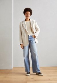 Beige liinapel blazer valge kudumisega sviitriga, koos helesiniste avarate pükste ja valgete tekstuuriga libisemisjalanõudega. Ükslane taust.