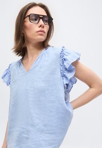 Γυναίκα με μαύρα γυαλιά και ανοιχτό μπλε μπλουζάκι με V λαιμόκοψη και βολάν στα μανίκια, κοιτάζοντας στο πλάι, με απλό λευκό φόντο.