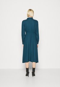 Tyrkysové šaty s dlouhým rukávem, s vypasovaným pasem, splývavou sukní a límečkem. Nosené s černými kotníkovými botami, vystavené na bílém pozadí.