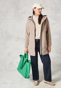 Veste imperméable beige avec capuche, associée à un pull crème, un jean foncé et des chaussures beiges. Tenue d'un grand sac fourre-tout vert.