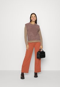 Sándo de textura mauve, sem mangas, sobre uma camisa de mangas longas creme com nervuras. Combinado com calças largas de cor ferrugem e uma bolsa preta.