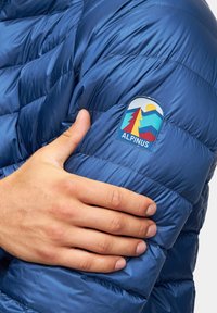 Niebieska kurtka puchowa z pikowanym wzorem, metalicznym połyskiem i haftowanym emblematem na lewym rękawie z motywami gór i słońca.