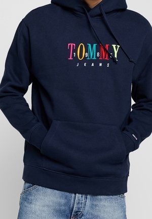 Sudadera de marina con colorido logo de "Tommy Jeans" en el pecho, bolsillo frontal y cordones, combinada con jeans de un azul claro.