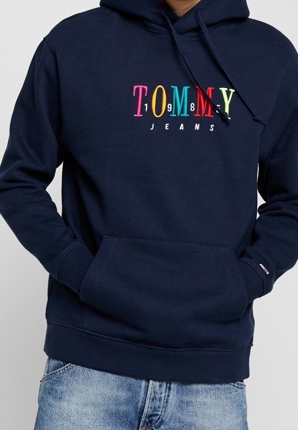 Sudadera de marina con colorido logo de "Tommy Jeans" en el pecho, bolsillo frontal y cordones, combinada con jeans de un azul claro.