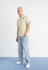 Camiseta de algodón beige con un logo verde, combinada con jeans de mezclilla azul claro. El modelo está de pie sobre una alfombra azul en un fondo minimalista.