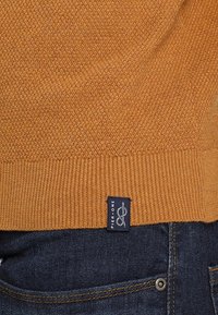 Närbild av en texturerad brun stickad tröja med en svart etikett som läser "Pier One" ovanför mörkblå denimjeans.