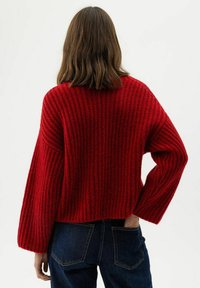 Persona con capelli castani lunghi fino alle spalle, che indossa un maglione a coste rosso e jeans blu scuro, in piedi con una mano nella tasca posteriore.