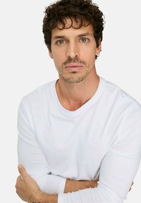 Witte shirt met lange mouwen, ronde halslijn, aansluitende mouwen en een subtiel logo op de borst. De achtergrond is effen wit.