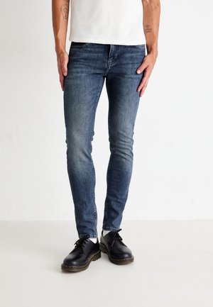 Jean en denim bleu skinny avec une finition délavée, porté avec des chaussures en cuir noires, présentant une coupe ajustée et un minimum d'usure.