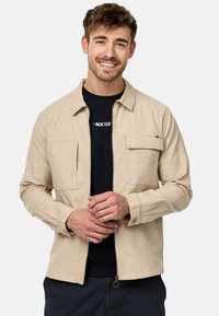 Veste beige zippée en tissu léger, dotée de deux poches avant. Le modèle porte un t-shirt noir en dessous.