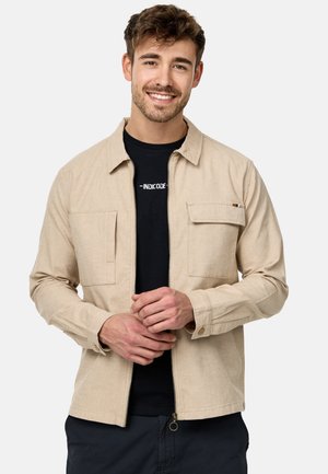 Beige, lynlåsjakke lavet af letvægtsstof, med to frontlommer. Modellen har en sort t-shirt på nedenunder.