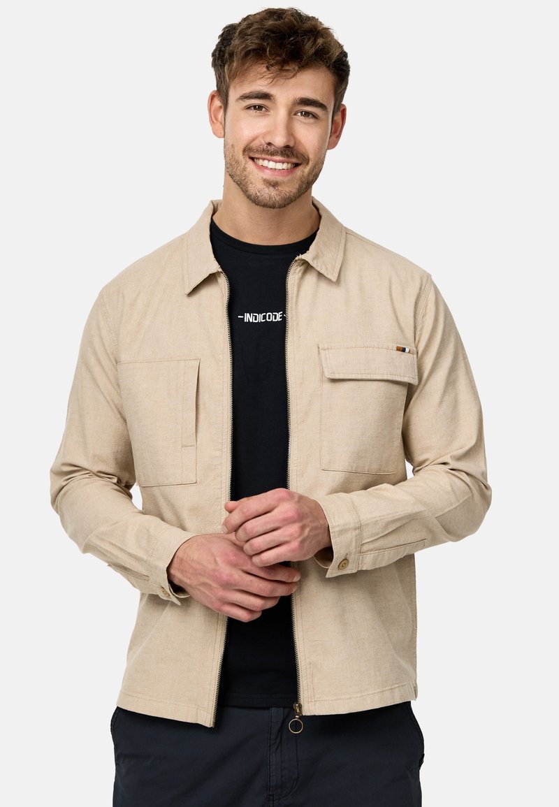 Veste beige zippée en tissu léger, dotée de deux poches avant. Le modèle porte un t-shirt noir en dessous.