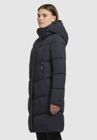 Polstret mørkeblå frakke med hætte, lynlåslomme på siden og knælangt design med definerede quiltede sektioner.